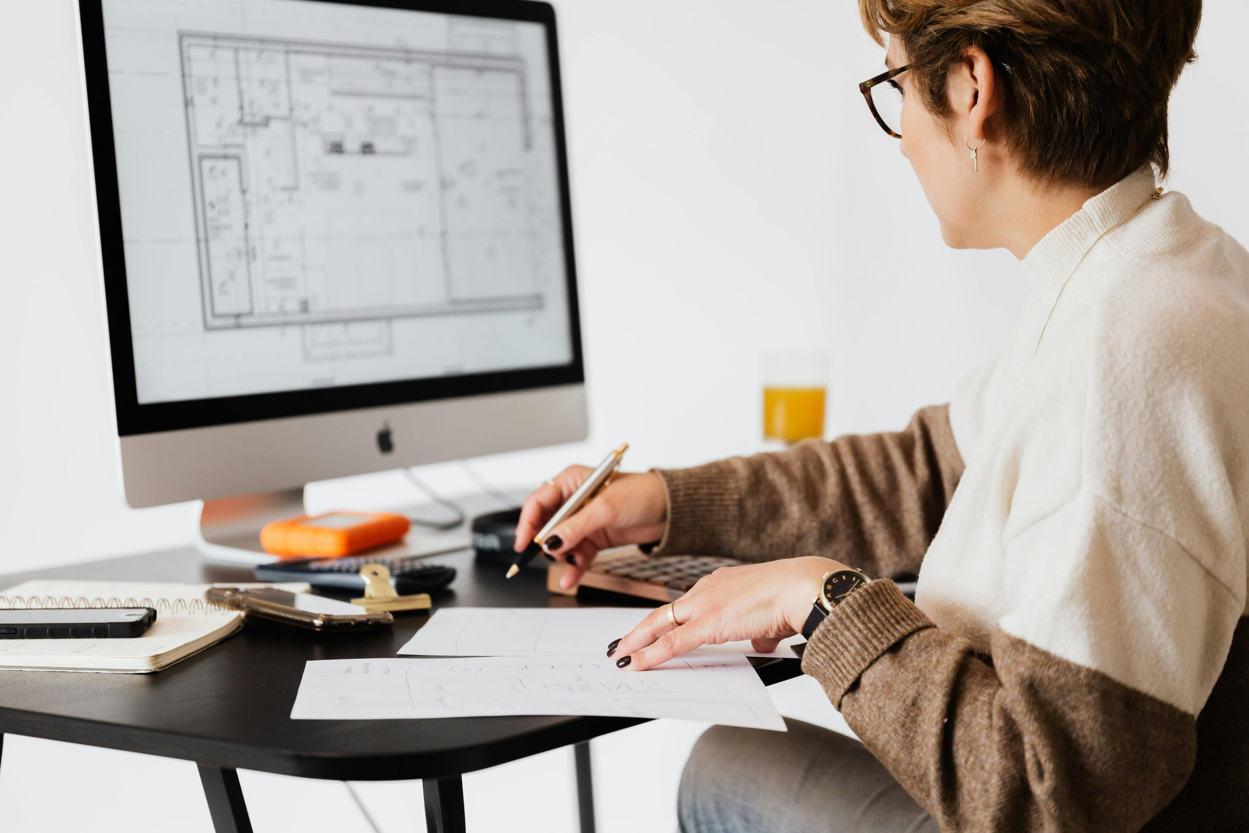 Professional analyzing technical blueprints on a computer screen while reviewing documents at a desk.